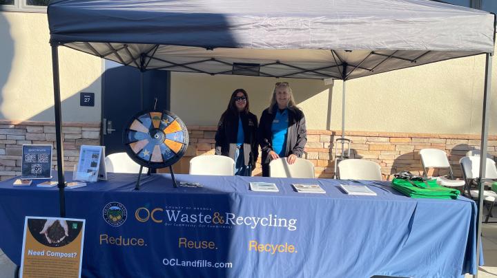 Two women standing behind a table with a tablecloth reading "OC Waste and Recycling"."