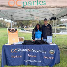 One woman and one man standing on grass behind a table with a tablecloth reading "OC Waste and Recycling""