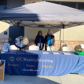 Two women standing behind a table with a tablecloth reading "OC Waste and Recycling"."