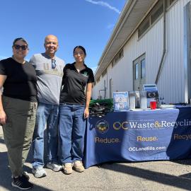 Three women and one man standing in front of a table with a tablecloth reading "OC Waste and Recycling"."