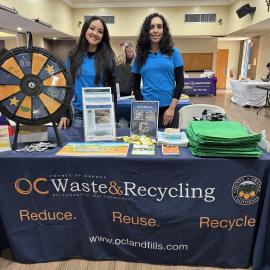 Two women standing behind a table with a tablecloth reading "OC Waste and Recycling"."